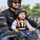 A happy toddler wearing a helmet and safety harness securely attached to an adult rider on a motorcycle.